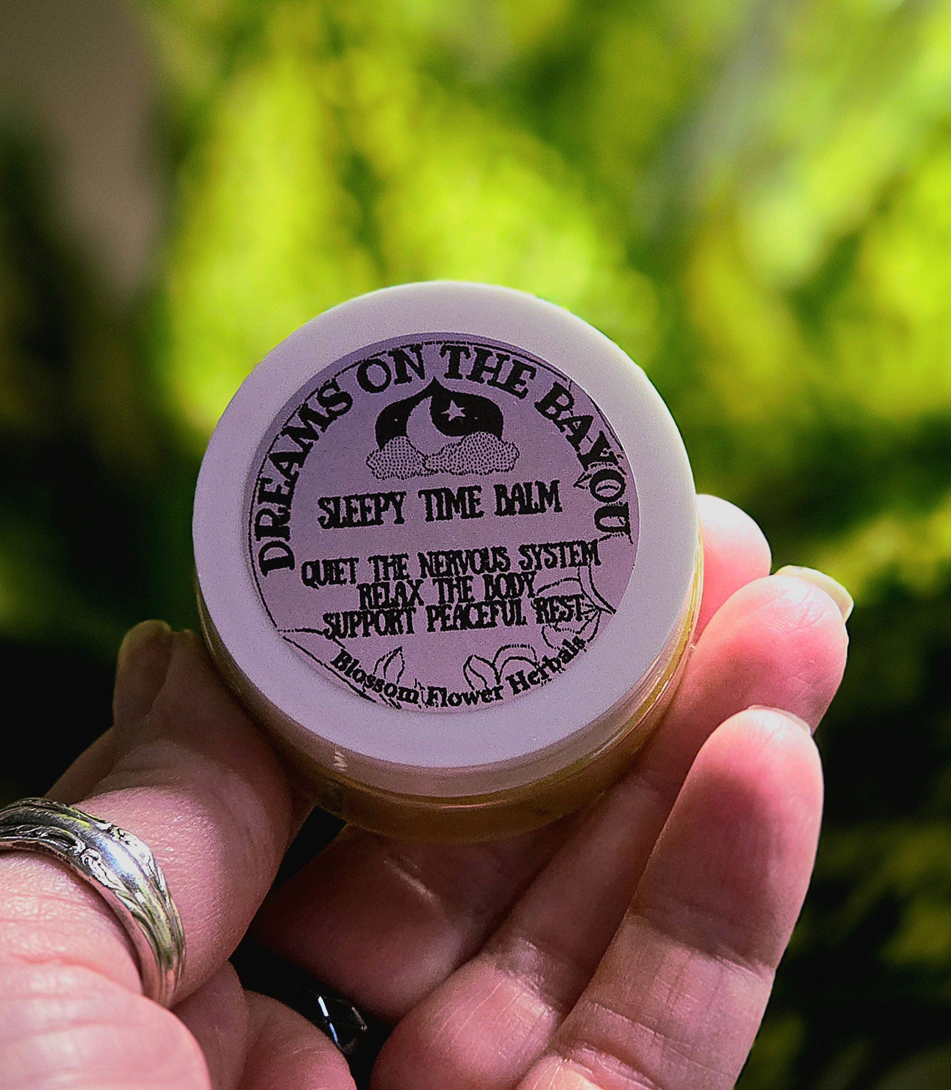 Hand holding a small container of 'Dreams on the Bayou' balm with a blurred green background